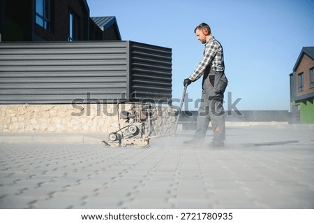 Landscaping worker using vibratory tamping machine plate compactor on a new patio driveway pavement to apply Polymeric sand to fill joints. Home garden and landscape renovation project background.