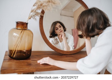 Languid Woman In A White Dress In Front Of The Mirror. Beautiful Brunette With Short Hair Indoors. From Behind.