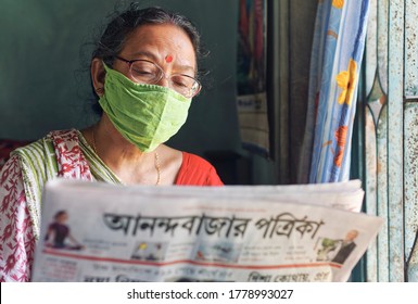 Lansdowne, Kolkata, 07/18/2020: A simple looking mature Indian housewife reading a bengali newspaper sitting beside room window, in morning. She is wearing a face mask.
