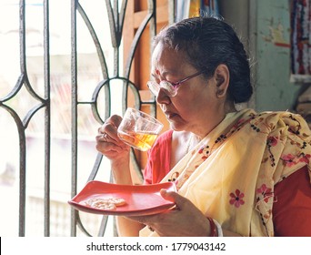 Lansdowne, Kolkata, 07/18/2020: A simple looking middle aged Indian housewife drinking her morning cup of golden tea liquor, sitting beside room window. She also has biscuits in her plate.