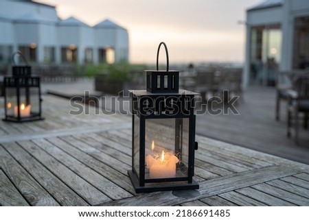 Lantern with a lit candle on an outdoor wooden table with sunset in the background for an outdoor party.