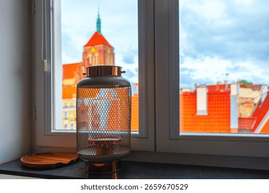 Lantern sits on a windowsill, casting a soft indoor contrast against backdrop of vibrant cityscape. Beyond glass, tiled rooftops and church rise into view, blending architectural charm of the city