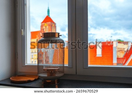 Lantern sits on a windowsill, casting a soft indoor contrast against backdrop of vibrant cityscape. Beyond glass, tiled rooftops and church rise into view, blending architectural charm of the city