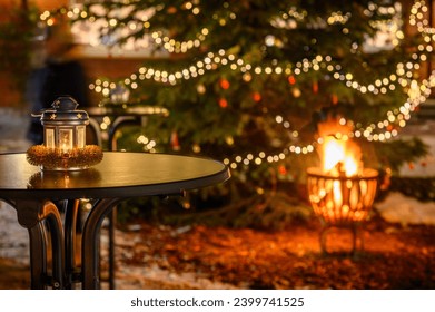 Lantern stands on the table next to a decorated Christmas tree, while a comforting fire flickers in the background. Lantern with festive touch illuminated with festive lights and warm glow of fire.