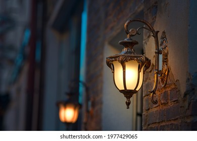 Lantern with yellow warm light on facade of building. Twilight on city street, building illumination. Retro lantern lighting, warm light glow. Street lights, illumination and vintage lantern - Shutterstock ID 1977429065