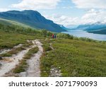 Lapland landscape with beautiful river Lulealven, snow capped mountain, birch tree and footpath of Kungsleden hiking trail with couple of hikers near Saltoluokta, north of Sweden. Summer blue sky