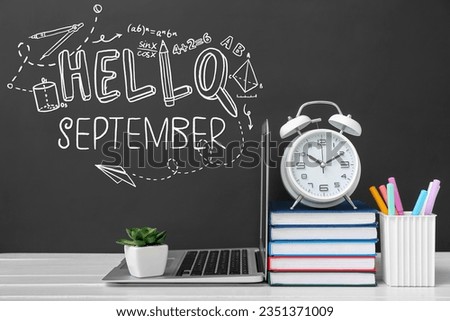 Laptop with alarm clock, books and stationery on table near chalkboard with written text HELLO SEPTEMBER
