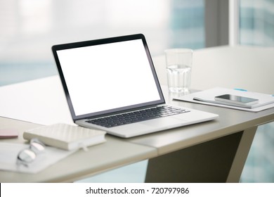 Laptop With Blank Screen On Desk In Modern Office Setting. Notepads, Glass Of Water, Reading Glasses And Phone On Table. Empty Copy Space For Business Ad, Corporate Website, Stock Investment Report.