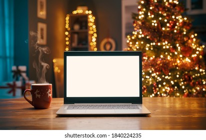 Laptop with blank screen on a desktop and Christmas tree with lights in the background - Powered by Shutterstock
