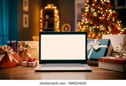 Laptop With Blank Screen On A Desktop At Home, Christmas Gifts And Decorated Tree In The Background