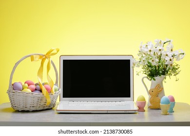 A laptop with a blank screen on a table festively decorated for the Easter holiday. Adorned working place with flowers and a basket with colorful eggs