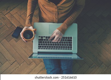 Laptop and coffee cup in girl's hands sitting on a wooden floor