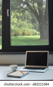 Laptop computer, notepad with pencil and cup of coffee on white desk in front of window at home office