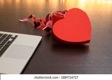 Laptop Computer And An Open Heart-shaped Gift Box On A Dark Wooden Table Against A Background Of Lights. Online Shopping And Chatting, Virtual Valentine's Day, Love In Time COVID-19 Concept.