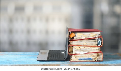 Laptop computer and stack of messy ringbinders with grungy business paperwork, bureaucracy,beat the clock,heavy workload concept. Blurred background.