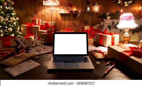 Laptop computer with white blank empty mock up screen on Merry Christmas table with presents gifts, decorated Xmas tree in Santa house background. Ecommerce website online shopping delivery ads.