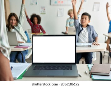 Laptop computer white blank empty mockup screen on teachers table with elementary junior children students raising hands in classroom background. Education software website technology ads concept.