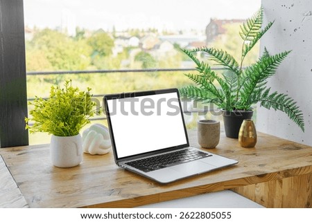 Laptop computer with white screen on wooden desk on wide window background. Office workspace, workplace in cafe or home. Mockup, template with copy space