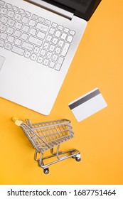 Laptop, credit card and small shopping trolley on yellow background, top view. Online shopping concept.