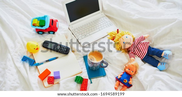 Laptop, cup of coffee, notebook, phone and\
different toys in bed on clean white linens. Freelance, distance\
learning or work from home with kids\
concept