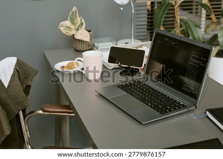 Laptop displaying code beside smartphone, mug, notebook, and snack on desk, showing remote software development workflow. Ideal for tech startup, coding, productivity, or home office content