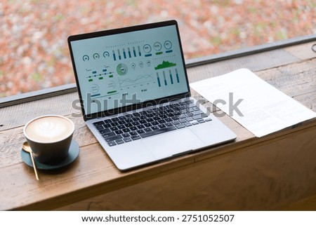 Laptop displaying data analytics charts on a wooden windowsill beside a coffee cup in autumn light