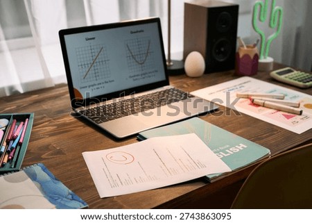 Laptop displaying math graphs on wooden desk surrounded by English workbook, graded assignment, calculator, and stationery suggesting online lesson or tutoring session setup