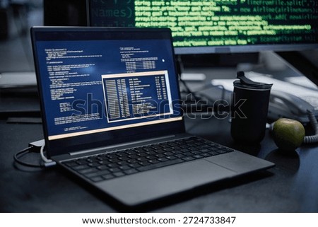 Laptop displaying programming code on screen sitting on desk with reusable cup and green apple nearby, computer monitor in background showing green text on black background