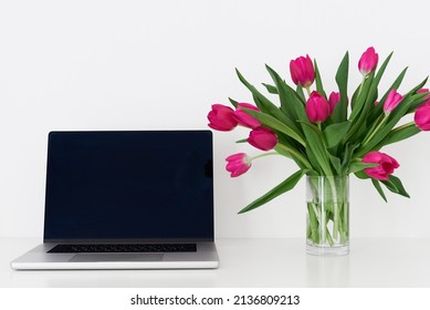 Laptop with empty black screen and bouquet of pink tulip flowers in a transparent vase on a white table. Working space. Home office