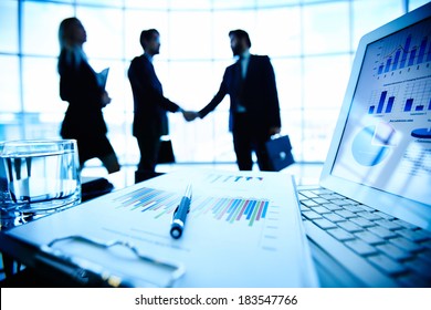 Laptop, financial document with pen and glass of water at workplace on background of three business partners making a deal