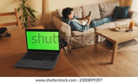 Laptop with green chromo key on the screen, mock-up screen, stands on table, in the background guy lying on sofa uses mobile phone