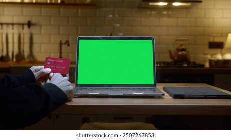 A laptop with a green screen on kitchen counter. A person holds a credit card to make an online purchase in the kitchen. Customizable content, online shopping, e-commerce, digital payments. - Powered by Shutterstock