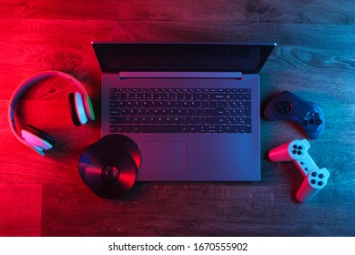 Laptop With Headphones, Gamepads, Cd Discs On A Wooden Floor. Blue Red Neon Light. Gradient Glow. Gaming Still Life. Top View