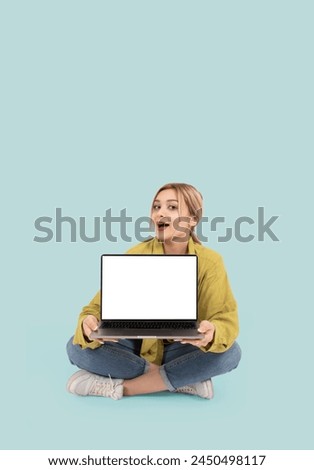 Laptop mock up, young smiling fun caucasian blonde woman hold show laptop mock up. Full body length girl sit floor legs crossed wear casual denim jean and shirt. Show empty blank screen pc computer.