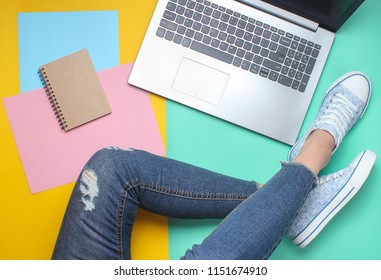 Laptop, notebook, female legs in jeans and sneakers on a colored pastel background, minimalism, flat lay style