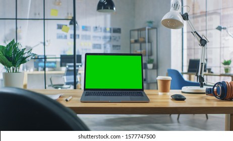 Laptop On The Desk In The Office Shows Green Mock-up Screen. In The Background Creative Modern Open Space Loft Office HUB With Professional Working