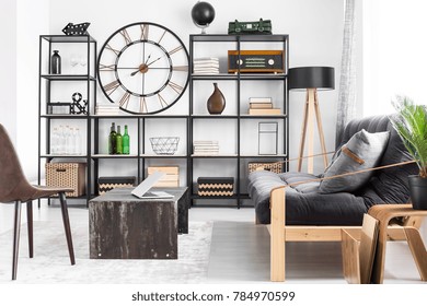Laptop on table and grey pillow on couch in man's living room interior design with metal clock on the wall between shelves