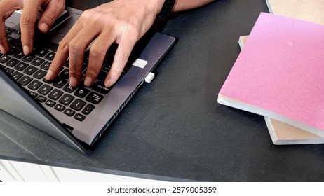 Laptop on a table with a stack of books next to someone's hands typing