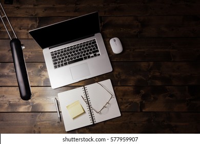 Laptop On Wooden Desk Viewed From Above. Notepad And Reading Glasses Next To Computer And A Desk Light Illuminating The Table.