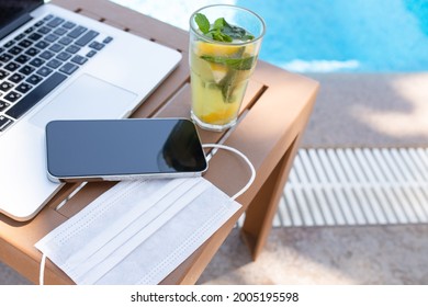 Laptop, Phone, Mojito Cocktail And Medical Mask On The Table By The Pool. Nobody. Concept Of New Rules, Masks In Public Places, Protection Against Coronavirus, Freelance, Vacation, Summer Work Place.