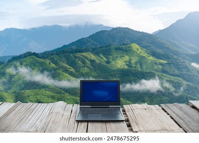 A laptop placed on a wooden deck with a breathtaking view of lush green mountains and misty clouds in the background. - Powered by Shutterstock