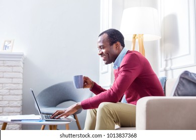 At the laptop. Positive joyful nice man pressing a button and looking at the laptop screen while holding a cup of tea