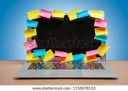 Laptop in reminders with colored stickers notes on the display frame on table with blue wall. Many unfulfilled tasks, deadline