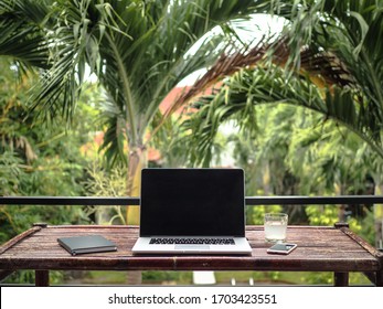 laptop of a remote digital nomad on a wooden bamboo table with notebook, mobile phone and glass in nature with a green tropical background with palm trees