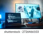 Laptop screen displaying programming code in foreground, large monitor in background showing surveillance footage of office with Caucasian young man and coworkers working at office desks