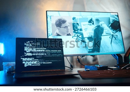 Laptop screen displaying programming code in foreground, large monitor in background showing surveillance footage of office with Caucasian young man and coworkers working at office desks