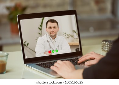 A laptop screen view over a woman's shoulder. A woman at an online doctor's appointment. A girl is discussing her health with a therapist in a white lab coat on a video call.