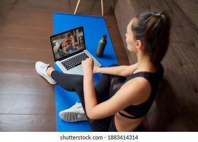 A Laptop Screen View Over A Woman's Shoulder. A Sporty Girl In A Tight Suit Is Watching A Workout Video On Laptop In Her Apartment. Woman Is Listening To A Coach During A Virtual Fitness Class At Home