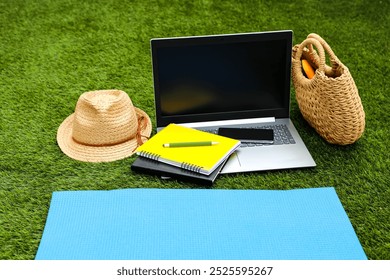 A laptop, a smartphone, a straw hat, a women's wicker bag, a beach mat and a notebook lie on the green grass. The Digital Nomads series. - Powered by Shutterstock