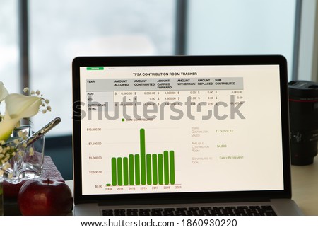 Laptop with spreadsheet of Savings account file, TFSA Contribution Tracker Document on desk. Yearly bar graph. Blurred window in background. White flowers, apple, and pen in container to the left. 